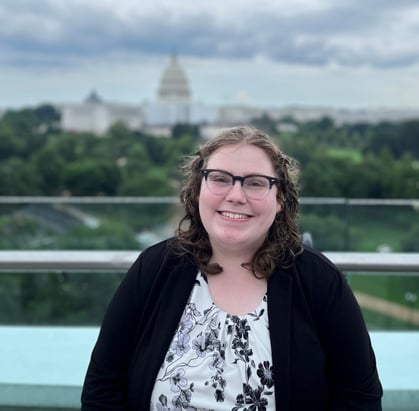 Elizabeth McCormick , a white woman with a floral shirt and glasses, smiles. The Capitol is in the distance behind her