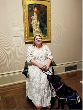 Katie, a white woman sitting in a power wheelchair wearing a long white dress with black print on it, smiles as she sits in front of a framed painting called Ophelia by Arthur Hughes. Katie’s black lab service dog, Petunia sits in front of her and is looking at Ophelia.