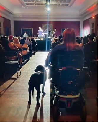 The back of Katie in her wheelchair and her service dog, Petunia, as they sit in the back of an audience all looking at a stage where John Legend sits at a piano giving an intimate concert at the Toledo Museum of Art.