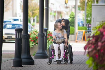 A couple kissing on the sidewalk. The woman is using a wheelchair and the man is leaning down behind her.