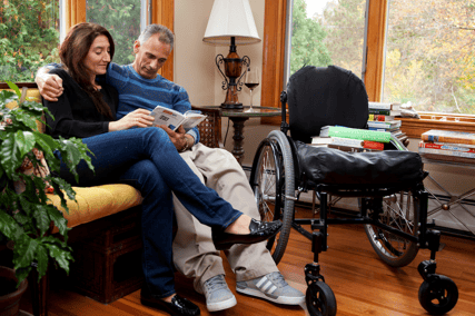 A couple sitting on a couch. The man's arm is around the woman and they are reading a book. There is a wheelchair next to them