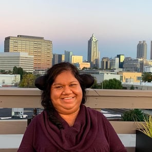 Liz, a woman with dark hair pulled in a twist to one side and an amber complexion, wears a maroon sweater on a rooftop with the Atlanta city skyline behind her. 