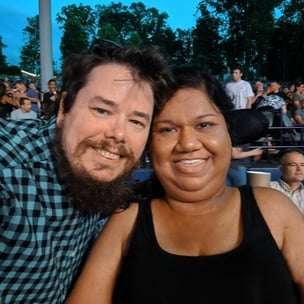 Liz, a woman with amber skin and dark hair pulled back, wears a black tank top as she smiles at the camera. Next to her is her husband Ben, a man with an ivory complexion and brown facial hair. He is also smiling while wearing a blue/black plaid shirt. 