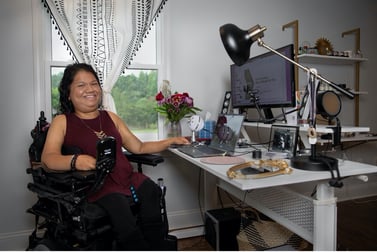 Liz sits in her power wheelchair in her well-organized home office which has a laptop and a separate monitor, a lamp, and other accessories. 