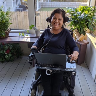 Liz sits in a power wheelchair with her laptop on a laptray. Her hair is pulled back. She smiles at the camera. She is in an enclosed porch with plants behind her. 