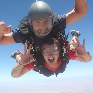 Dr. Toni Saia skydiving with an exuberant look on her face. She is attached to a skydiving instructor and is mid-air. 
