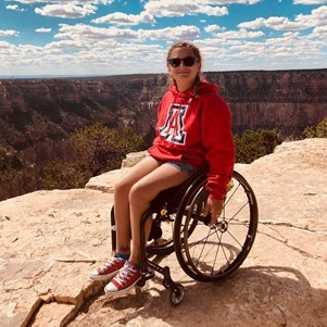 Dr. Toni Saia sits in her wheelchair, atop a canyon.  She is wearing a red hoodie with a big A on it and sunglasses. 