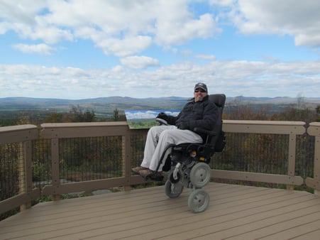 Man using a wheelchair on a scenic overlook. He is wearing a hat, sunglasses, and a coat.