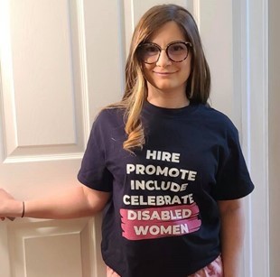 Dr. Toni Saia, a pale woman with long blonde hair, smiles at the camera. She wears a shirt that reads “Hire, promote, include, celebrate Disabled Women”