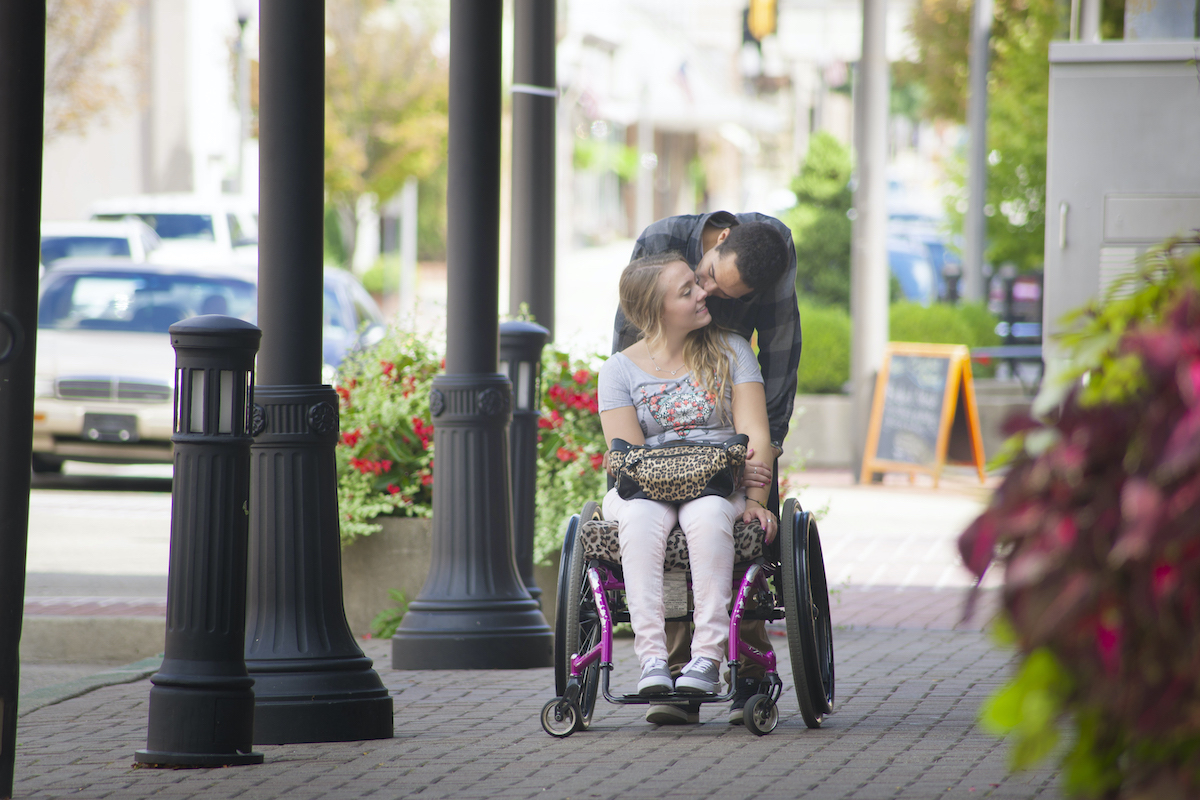 A couple kissing on the sidewalk. The woman is using a wheelchair and the man is leaning down behind her.