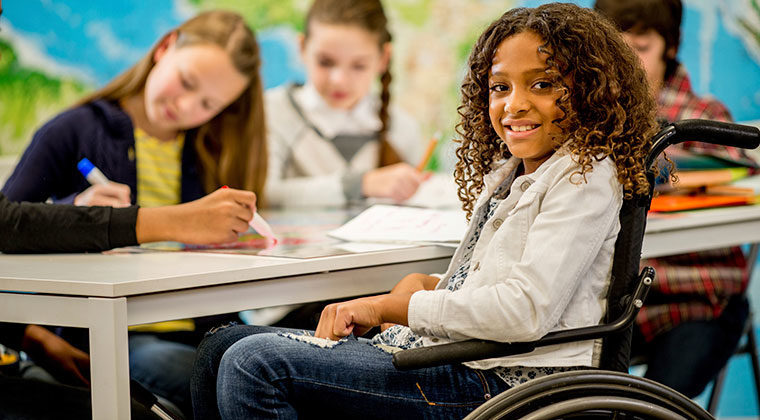 young girl with curly brown hair smiles while sitting in manual chair (foreground). Other students working (background)