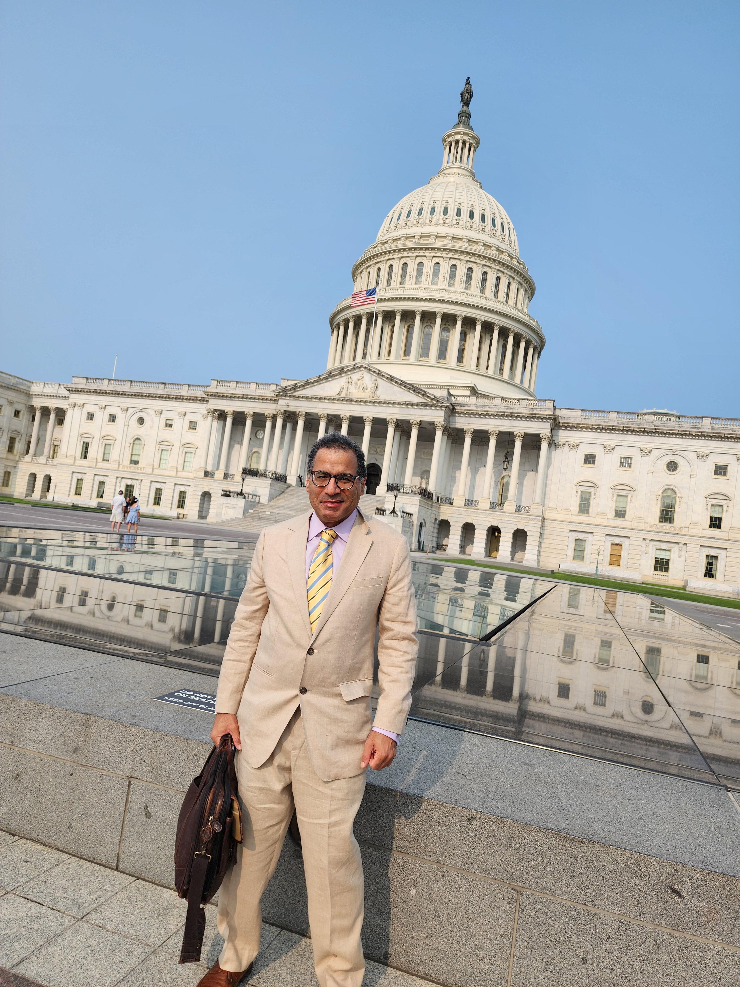 Marc, a light skinned Black man, standing in front of the United States Capitol Building in Washington, D.C. He is dressed formally in a beige suit with a white shirt and a yellow-striped tie. He is wearing glasses and has short dark hair. He is holding a dark brown leather bag in his left hand and is standing on what appears to be a reflective surface, as the Capitol building is partially reflected in front of him. 